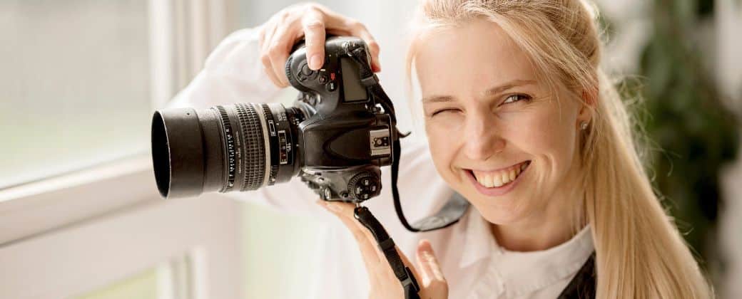 בחורה בלונדינית עם שיער אסוף מחזיקה מצלמה, קורצת עם עין אחת ומסבירה לגבי איך מצלמים מקצועי – לימודי סטודיו שמוכיחים את עצמם