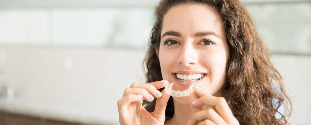 אישה מתולתלת עם שיער חום מחזיקה פלטה שקופה ותוהה למה יותר מבוגרים בוחרים היום ביישור שיניים שקוף.